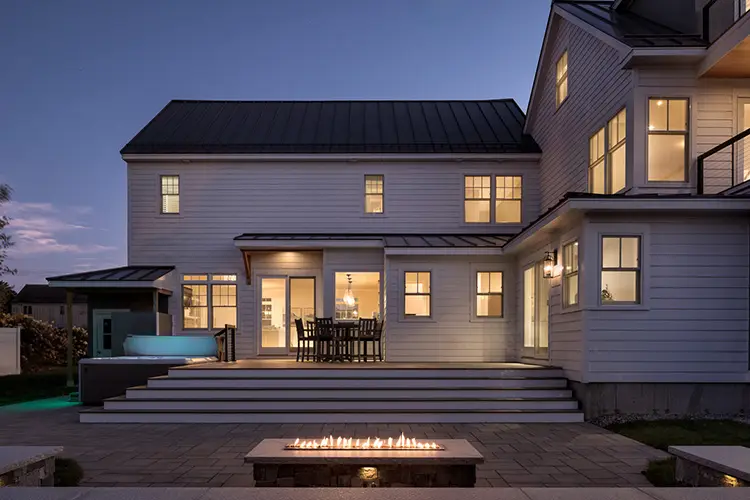 Driftwood Lane beach house at dusk with lit windows, stone patio and fire table in Pine Point, Scarborough, Maine